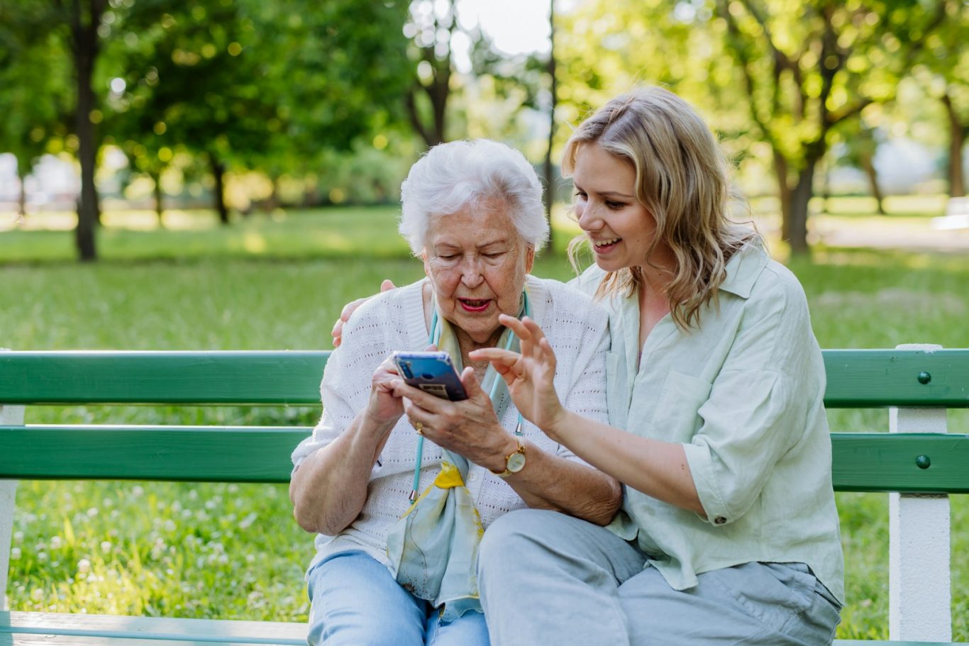 Smartphoneerklärung Eine ältere Frau und eine jüngere Frau sitzen auf einer Bank und schauen zusammen auf ein Smartphone.