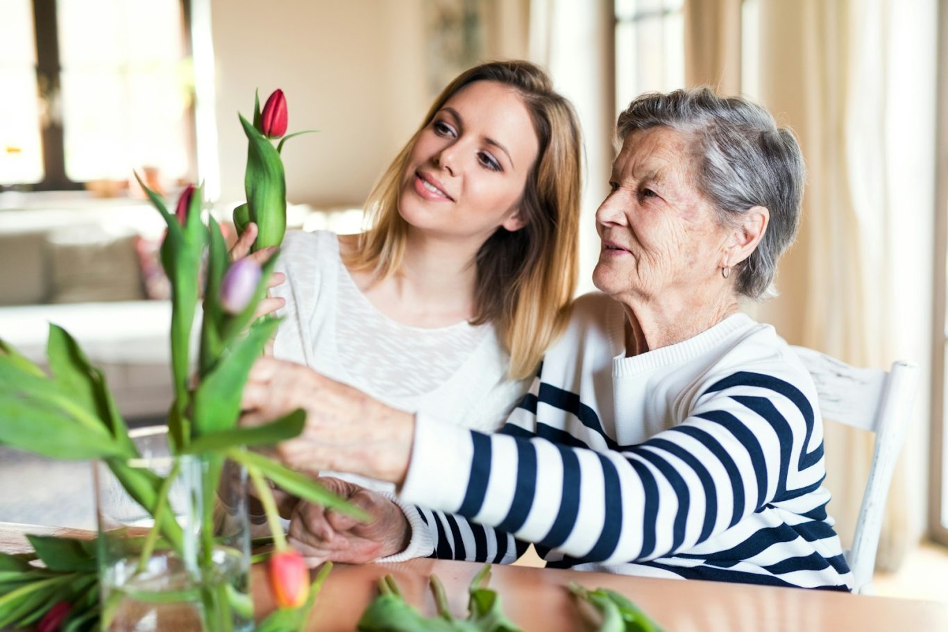 Seniorenbetreuung Junge Frau arrangiert gemeinsam mit älterer Dame Blumen in einer Vase.