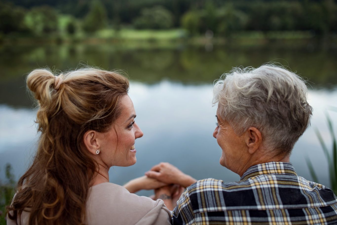 Spaziergang Junge Frau und ältere Dame sitzen am Ufer eines ruhigen Sees, schauen sich an.